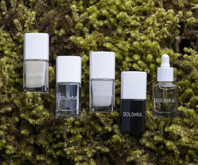 Fünf Dolomia Nagelpflege-Produkte in eckigen Glasflaschen und einer Pipettenflasche, angeordnet auf grünem Moos, Draufsicht.